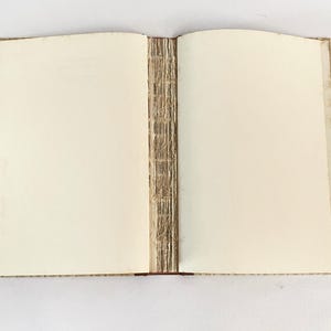 May include: An open, handmade journal with blank, cream-colored pages. The binding is visible, showing the hand-stitched spine. The cover edges have a textured, patterned design. The book is lying open, ready for writing or sketching.