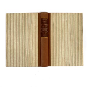 May include: A vintage book titled "Three Plays of Henrik Ibsen." The book has a brown leather spine with gold lettering and a beige cover with vertical striped patterns. The book's title and author are visible.