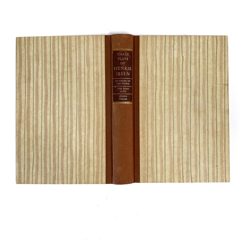 May include: A vintage book titled "Three Plays of Henrik Ibsen." The book has a brown leather spine with gold lettering and a beige cover with vertical striped patterns. The book's title and author are visible.
