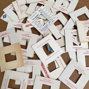 May include: A collection of square white cardboard photographic slide mounts scattered in a pile. Each mount features a central square cutout and printed text in red or blue ink, including brand names like Anscochrome and Agfachrome.