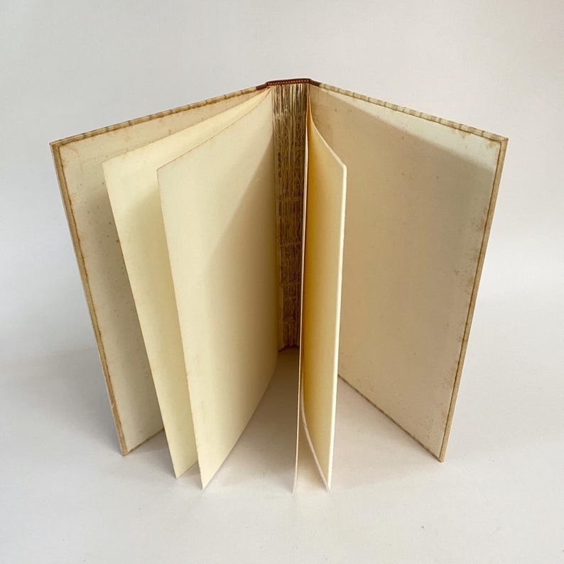 May include: An open, antique book with aged, cream-colored pages. The book's spine is visible, and the pages are slightly fanned out, revealing their edges. The cover appears to be a light tan color, with a worn appearance.