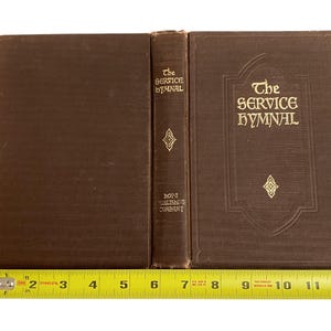 May include: A brown hardcover book titled "The Service Hymnal" with gold lettering and decorative elements. The spine reads "The Service Hymnal" and "Hope Publishing Company". The book is measured with a yellow tape measure.