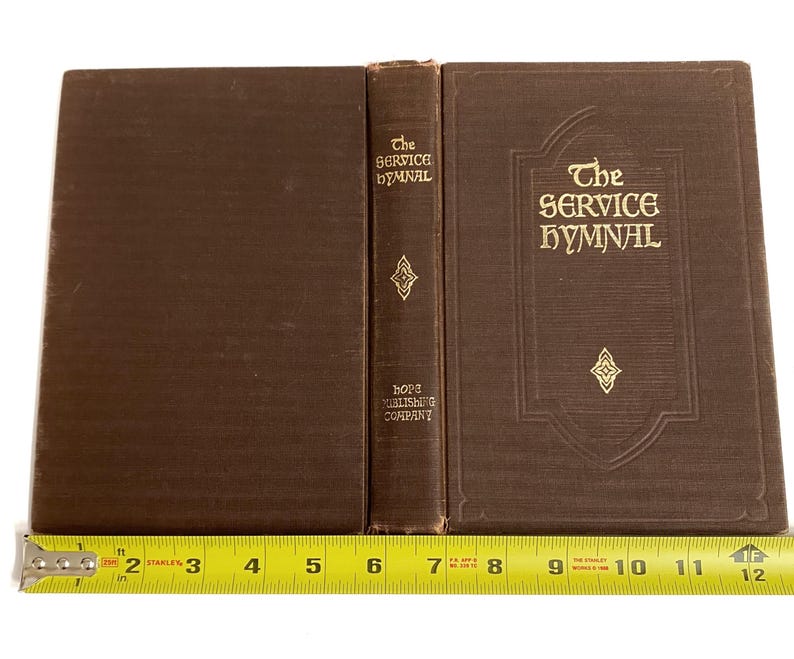 May include: A brown hardcover book titled "The Service Hymnal" with gold lettering and decorative elements. The spine reads "The Service Hymnal" and "Hope Publishing Company". The book is measured with a yellow tape measure.