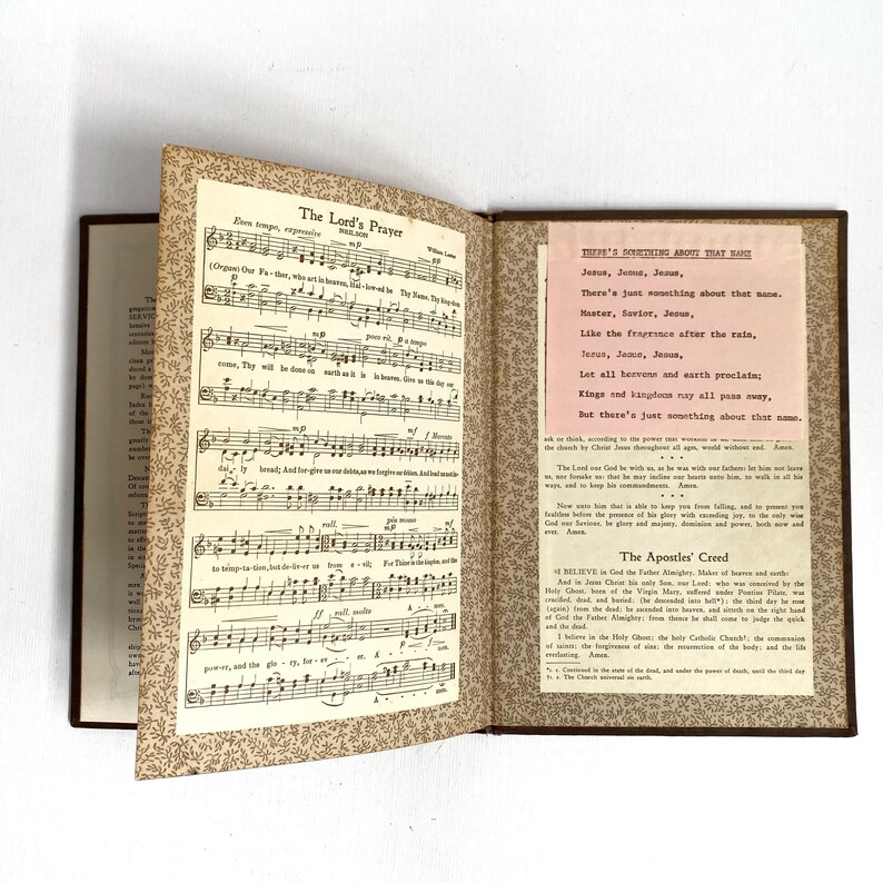 May include: An open, vintage book with brown covers and gold-toned patterned endpapers. The left page displays sheet music for "The Lord's Prayer." The right page features a pink insert with the text "There's Something About That Name" and "The Apostles' Creed."