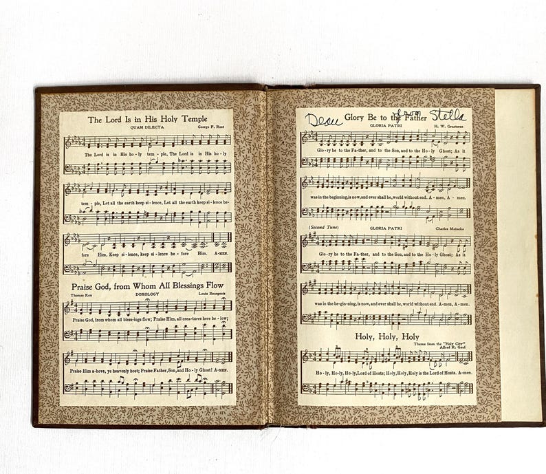 May include: An open book of sheet music with brown covers and decorative patterned endpapers. The left page features the title "The Lord Is in His Holy Temple" and "Praise God, from Whom All Blessings Flow." The right page is titled "Glory Be to the Father" and "Holy, Holy, Holy."