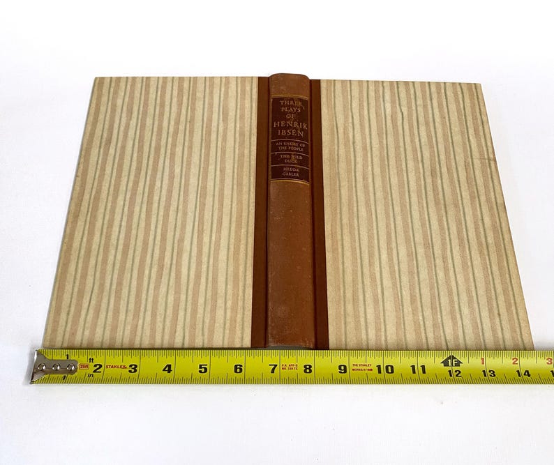 May include: A vintage book titled "Three Plays of Henrik Ibsen" with a brown leather spine and tan striped covers. A yellow measuring tape is placed below the book, showing measurements in inches.