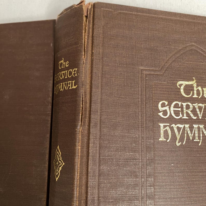 May include: Close-up of two antique brown books. The spine of one book displays the title "The Service Hymnal" in gold lettering. The other book's spine is visible, with the title partially visible. The books have a textured appearance.