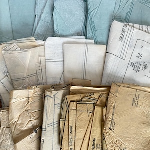 May include: A collection of folded sewing pattern pieces made of translucent white and tan paper. The pieces feature printed black lines, markings, and text including 'FRONT FACING B', 'FLAP', and '7575 MISS SIZES 8 AND 10'.