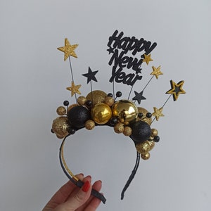 Könnte beinhalten: Ein festliches Neujahrs-Stirnband mit goldenen und schwarzen Dekorationen. Das Stirnband ist mit goldenen und schwarzen Glitzerkugeln, Sternen und dem Schriftzug "Happy New Year" verziert. Ein lustiges Accessoire zum Feiern des neuen Jahres.