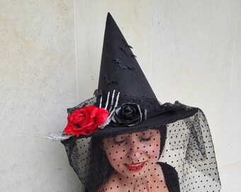 May include: A black witch hat with a tall, pointed crown and a wide brim. The hat is decorated with a red rose, a black rose, and a skeleton hand. A black veil with polka dots drapes over the face.