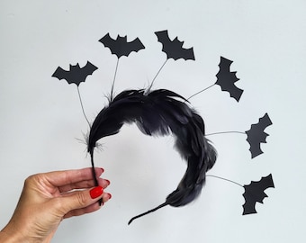 May include: A black Halloween headband featuring a central arrangement of black feathers. Six black bat cutouts are attached to thin wires, radiating outwards from the feather base. The headband is held against a white background.