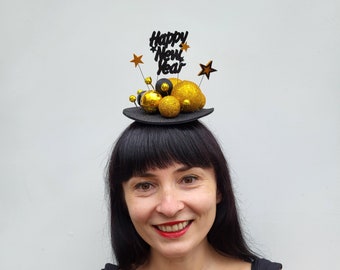 May include: A black mini top hat decorated with gold and black ornaments, gold stars, and the words "Happy New Year". The hat is worn by a person with black hair and red lipstick.
