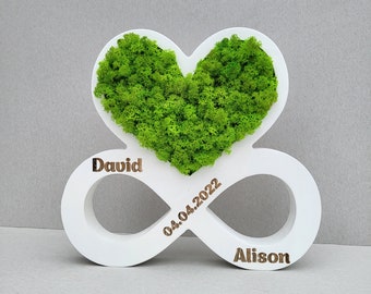 May include: White wooden infinity symbol with a heart shape in the center filled with green moss. The infinity symbol is engraved with the names "David" and "Alison" and the date "04.04.2022".