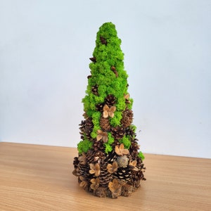Könnte beinhalten: Ein dekorativer, kegelförmiger Baum aus grünem Moos, Tannenzapfen und getrockneten Samenkapseln. Die Basis ist eine natürliche Holzscheibe. Eine natürliche, rustikale Dekoration.