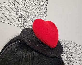 May include: A black fascinator with a red heart-shaped accent and a delicate black net veil. The base is a small, round, black disc with glitter. The headpiece is worn on the head.