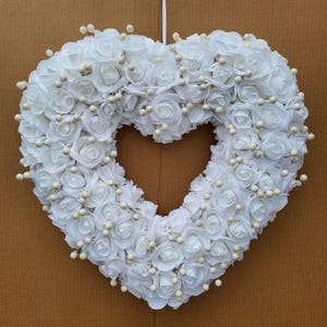 以下が含まれることがあります： 白いハート型のリースは、造花と小さな白いビーズでできています。リースは茶色の壁にかけられています。