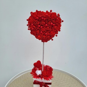 May include: A decorative floral arrangement with a heart-shaped top made of red roses and berries. The heart sits atop a white stem, with a base of red feathers, roses, and a small heart. A red ribbon adorns the white square base.
