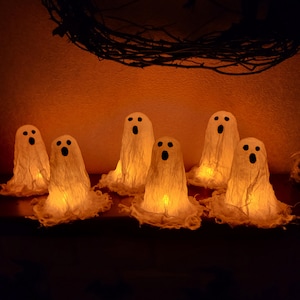 Op de afbeelding: Een verzameling van zes kleine, spookvormige decoraties gemaakt van dunne, doorschijnende stof. Elk spookje heeft een handgetekend zwart gezicht met twee ronde ogen en een ovale mond, verlicht van binnenuit door een warm, oranje schijnsel.