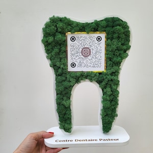 Op de afbeelding: Een wit, tandvormig decoratief stuk bedekt met groen mos. Een QR-code met een Instagram-logo is gecentreerd op het mos. De basis luidt "Centre Dentaire Pasteur".