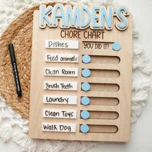 May include: A light wood chore chart with the name Kamden's in blue letters at the top. It features seven white horizontal slots for tasks including Dishes, Feed animals, Clean Room, Brush Teeth, Laundry, Clean Toys, and Walk Dog, with blue sliding markers and a black dry erase marker.