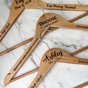 May include: Four wooden hangers with personalized text. The hangers are engraved with names, school names, and the year "2024". The text on the hangers reads "Kaya Class of 2024", "Mortimer Jordan High School", "Brodie Class of 2024", "Hayden High School", "Ashley Class of 2024", and "Keep Moving Forward".