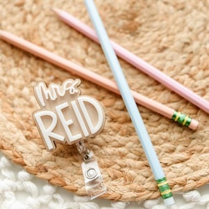 May include: A badge reel with the text "Mrs. REID" in white lettering, along with three pencils in pastel colors: pink, peach, and light blue. The pencils are on a woven, circular mat.