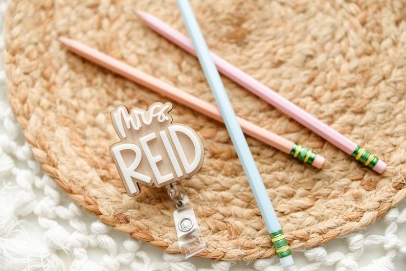 May include: A badge reel with the text "Mrs. REID" in white lettering, along with three pencils in pastel colors: pink, peach, and light blue. The pencils are on a woven, circular mat.