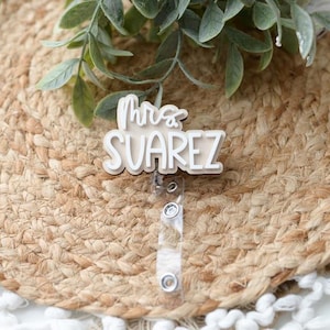 May include: A white badge reel with the text "Mrs. Suarez" in a script font. The badge is on a woven, circular placemat with green leaves. The reel has a clear plastic strap and a metal clip.