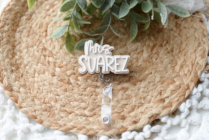 May include: A white badge reel with the text "Mrs. Suarez" in a script font. The badge is on a woven, circular placemat with green leaves. The reel has a clear plastic strap and a metal clip.