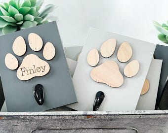 May include: Two wooden paw print hooks, one gray and one white, with a black hook on each. The gray hook has the name "Finley" engraved on it.
