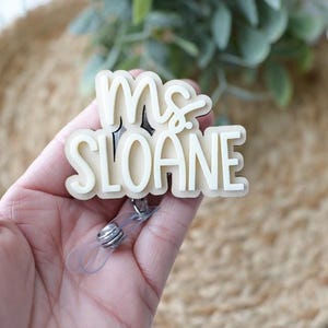 May include: A cream-colored badge reel with the text "Ms. Sloane" in a layered design. The badge reel has a clear plastic retractable cord and a metal clip. The background is blurred, with a natural, neutral color palette.