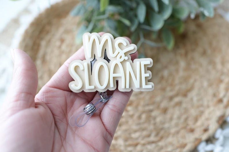 May include: A cream-colored badge reel with the text "Ms. Sloane" in a layered design. The badge reel has a clear plastic retractable cord and a metal clip. The background is blurred, with a natural, neutral color palette.