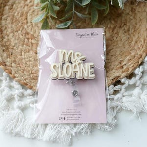 May include: A badge reel with the name "Ms. Sloane" in a layered, off-white and beige design. The badge is packaged on a light pink card with the text "Forged on Main". The background includes a woven mat and white fringed fabric.