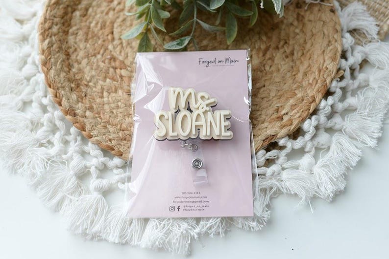 May include: A badge reel with the name "Ms. Sloane" in a layered, off-white and beige design. The badge is packaged on a light pink card with the text "Forged on Main". The background includes a woven mat and white fringed fabric.