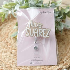 May include: A badge reel with the name "Mrs. Suarez" in a layered design. The badge reel is packaged on a pink card with contact information and social media handles. The background includes greenery and a woven surface.