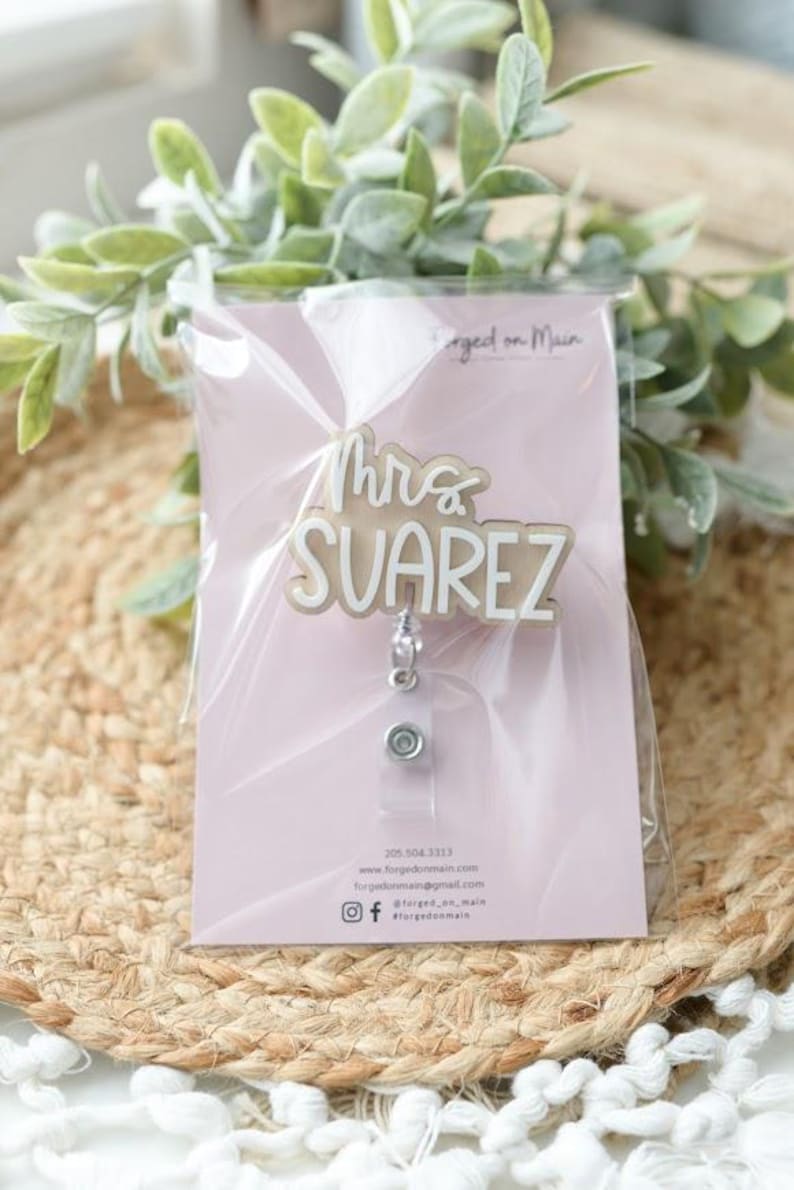 May include: A badge reel with the name "Mrs. Suarez" in a layered design. The badge reel is packaged on a pink card with contact information and social media handles. The background includes greenery and a woven surface.