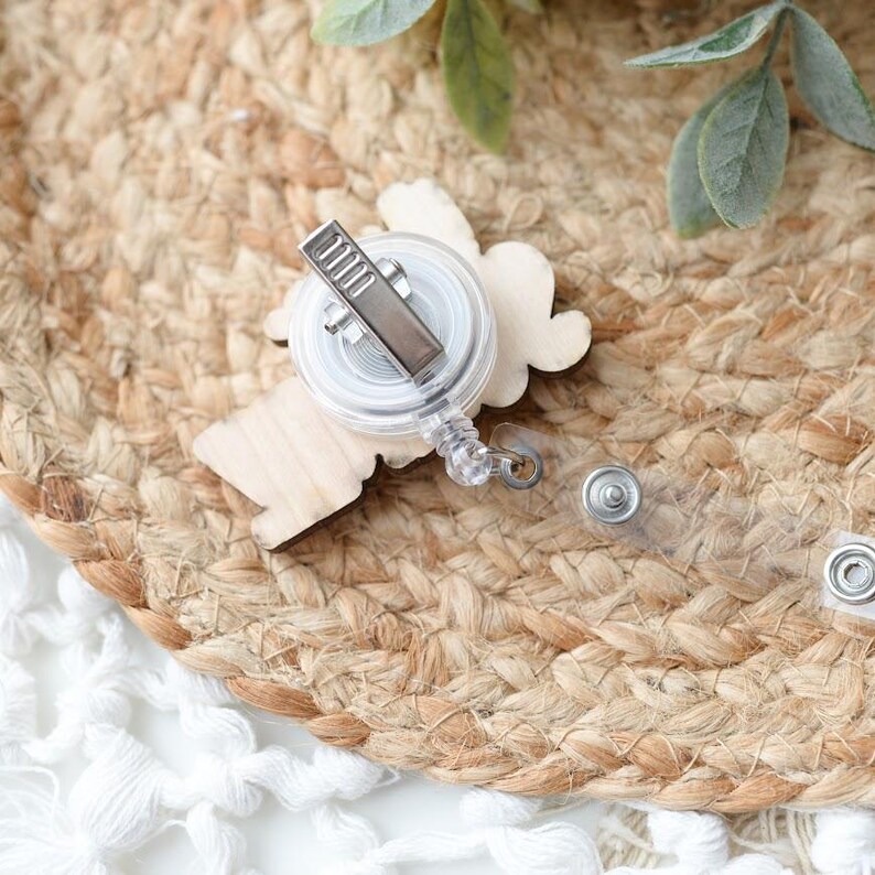 May include: A wooden badge reel with a light-colored, cloud-shaped design. The reel has a clear plastic casing, a silver metal clip, and a retractable cord. The badge reel is on a woven, tan surface with a white lace trim.
