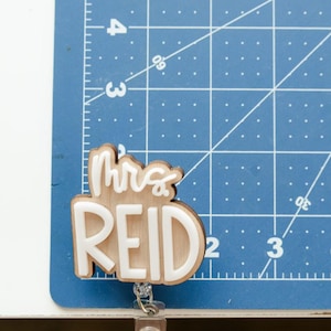 May include: A wooden name badge with the text "Mrs REID" in white lettering. The badge is attached to a retractable reel. The background is a blue cutting mat with white grid lines and measurements in inches.