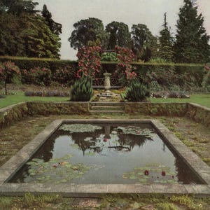 May include: A rectangular pond with a stone border and steps leading up to a small fountain in the middle of a garden. The pond is surrounded by lush green grass and trees.