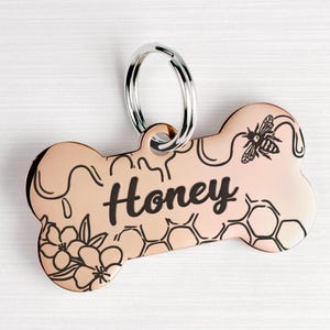 May include: A rose gold bone-shaped dog tag with a black engraved design of honeycombs, flowers, and a bee. The tag is personalised with the name "Honey".