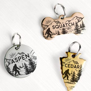 Op de afbeelding: Drie metalen hondenpenningen met berg- en Bigfoot-ontwerpen. Eén is rond met "ASPEN", een andere is botvormig met "SQUATCH" en de derde is pijlpunvormig met "CEDAR". Elke penning heeft een silhouet van Bigfoot en een bos.