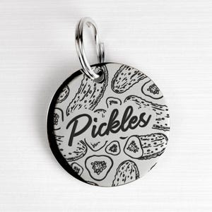 May include: A round, silver-colored pet tag with a black and white pickle design. The word "Pickles" is written in a cursive font across the center of the tag. A silver ring is attached at the top for easy attachment to a pet's collar.