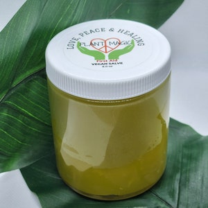 May include: A clear jar with a white lid, containing a green salve. The lid has a label with the text "LOVE, PEACE & HEALING" and "PLANT MAGIC". The jar is sitting on a large green leaf. The label also says "First Aid VEGAN SALVE 8.0 oz".