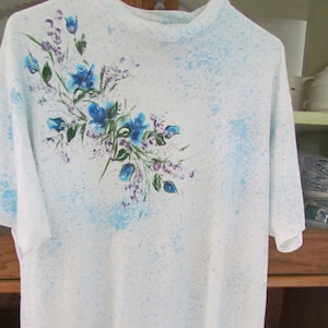 May include: White t-shirt with a mock turtleneck collar. The shirt features a floral design of blue flowers and green leaves on the left side. The shirt has a light blue speckled pattern throughout.