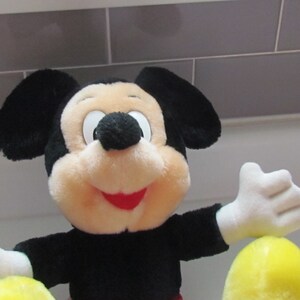 May include: A plush Mickey Mouse toy with black ears, a black body, and a cream-colored face. The toy has white gloves and a red mouth. The toy is holding a yellow object. The background is a light gray tiled wall.