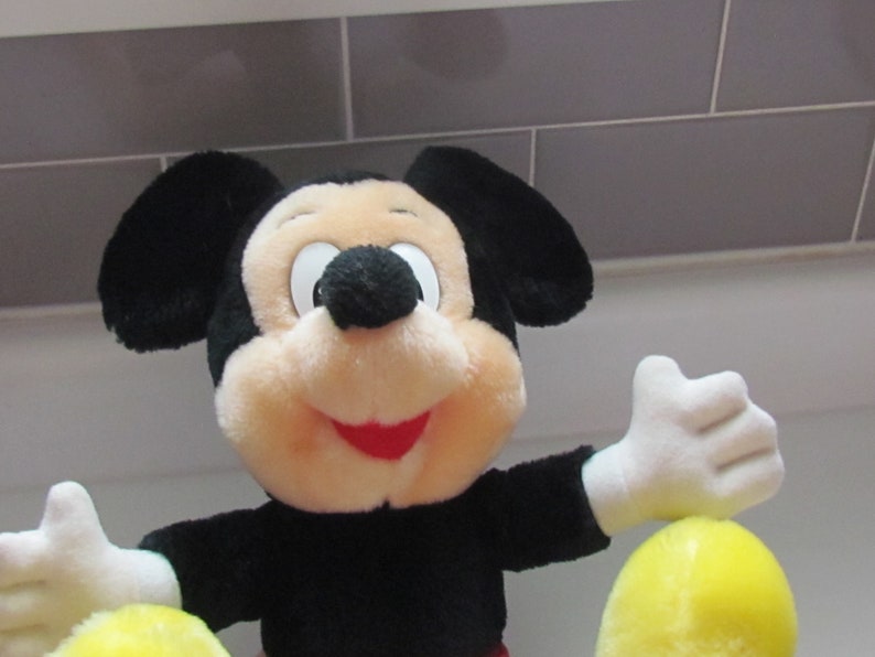 May include: A plush Mickey Mouse toy with black ears, a black body, and a cream-colored face. The toy has white gloves and a red mouth. The toy is holding a yellow object. The background is a light gray tiled wall.