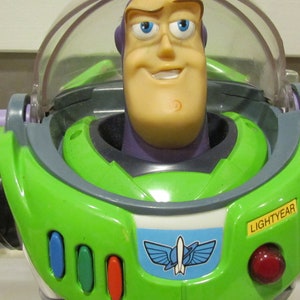 May include: A close-up of a Buzz Lightyear action figure. The toy has a green and white spacesuit with a clear helmet. The face is tan with blue eyes and a smile. The word "LIGHTYEAR" is visible on the chest.