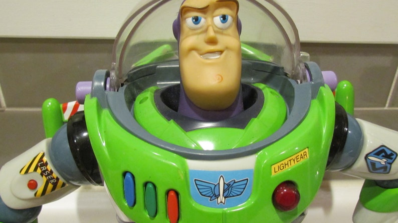 May include: A close-up of a Buzz Lightyear action figure. The toy has a green and white spacesuit with a clear helmet. The face is tan with blue eyes and a smile. The word "LIGHTYEAR" is visible on the chest.