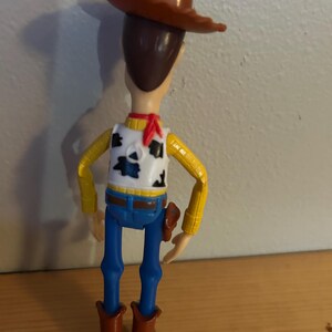 May include: A Woody action figure, viewed from the back, wearing a brown cowboy hat, a white vest with black cow spots, a yellow shirt, blue jeans, and brown boots. A red scarf is around the neck. A toy gun is in a holster.