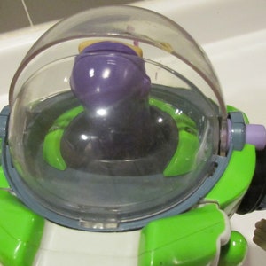 May include: A close-up of a Buzz Lightyear action figure. The toy has a clear plastic helmet, a purple head, and a green and white body. The figure is a popular character from the Toy Story franchise.
